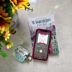 a picture of a phone opened to an audiobook for kids. the phone is on top of a copy of the vanderbeekers of 141st street and a pair of airpods and a pot of sunflowers are nearby